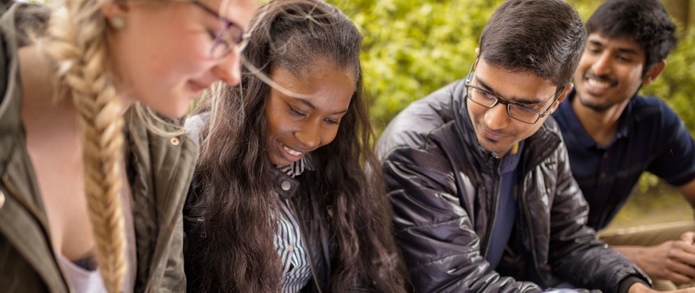 Deutsche und internationale Studierende sehen sich auf dem Campus sitzend gemeinsam etwas an, das nicht im Bild ist.