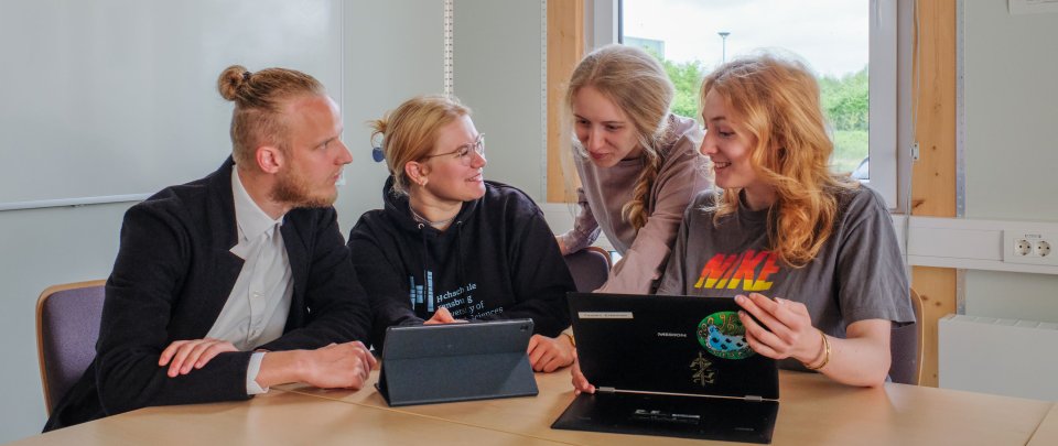 Eine kleine Gruppe Studierender sitzt nebeneinander und schaut auf einen Laptop. Eine Studentin zeigt den anderen etwas.