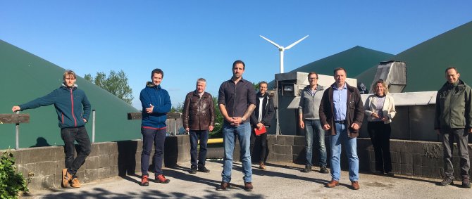 Eine Gruppe Menschen posiert draußen, der Himmel ist strahlend blau. Zu erkennen sind außerdem ein Windrad, Silos und Bäume.