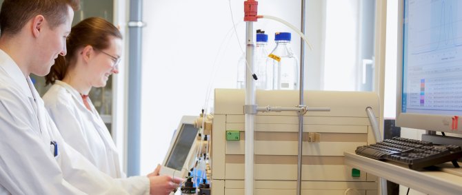 Zwei Studierende im Labor. Sie tragen Kittel und schauen auf ein Gerät und Bildschirm vor ihnen.