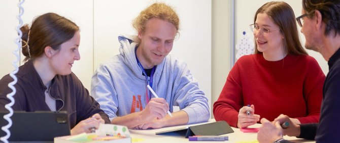 Studierende und ein Dozent stehen um einen Tisch. Sie halten Stifte, einer macht Notizen auf dem Tablet.