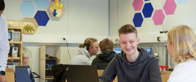 Studierende in einem freundlichen Arbeitsraum, sie sitzen an Tischen, halten Platinen in der Hand und reden.
