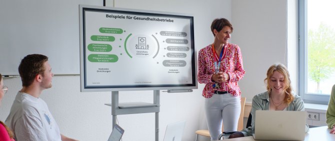 Seminarszene: Eine Dozentin steht vor einem Whiteboard, am Tisch sitzt eine Gruppe von vier Studierenden, die lächelnd auf ihre Laptops oder nach vorn zum Whiteboard schauen. Auf dem Whiteboard ist eine Grafik mit der Überschrift "Beispiele für Gesundheitsbetriebe" zu sehen.