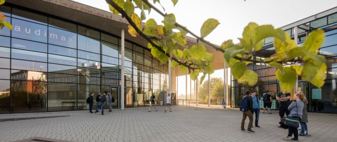 Studierende vor dem audimax. Es ist sonnig, am vorderen Bildrand ist der Ast eines Baumes zu erkennen.