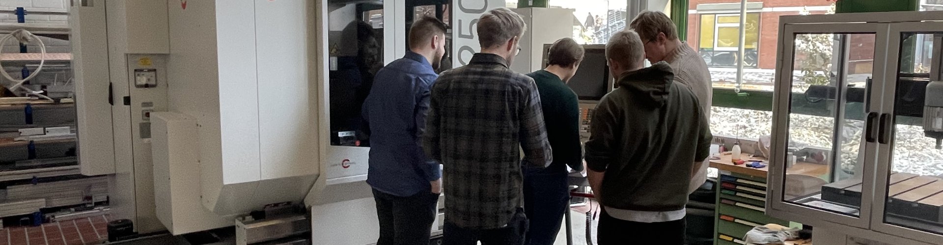 Studierende stehen vor eine 5-Achsfräsmaschinen und analysieren den Ablauf eine Umrüstvorganges.